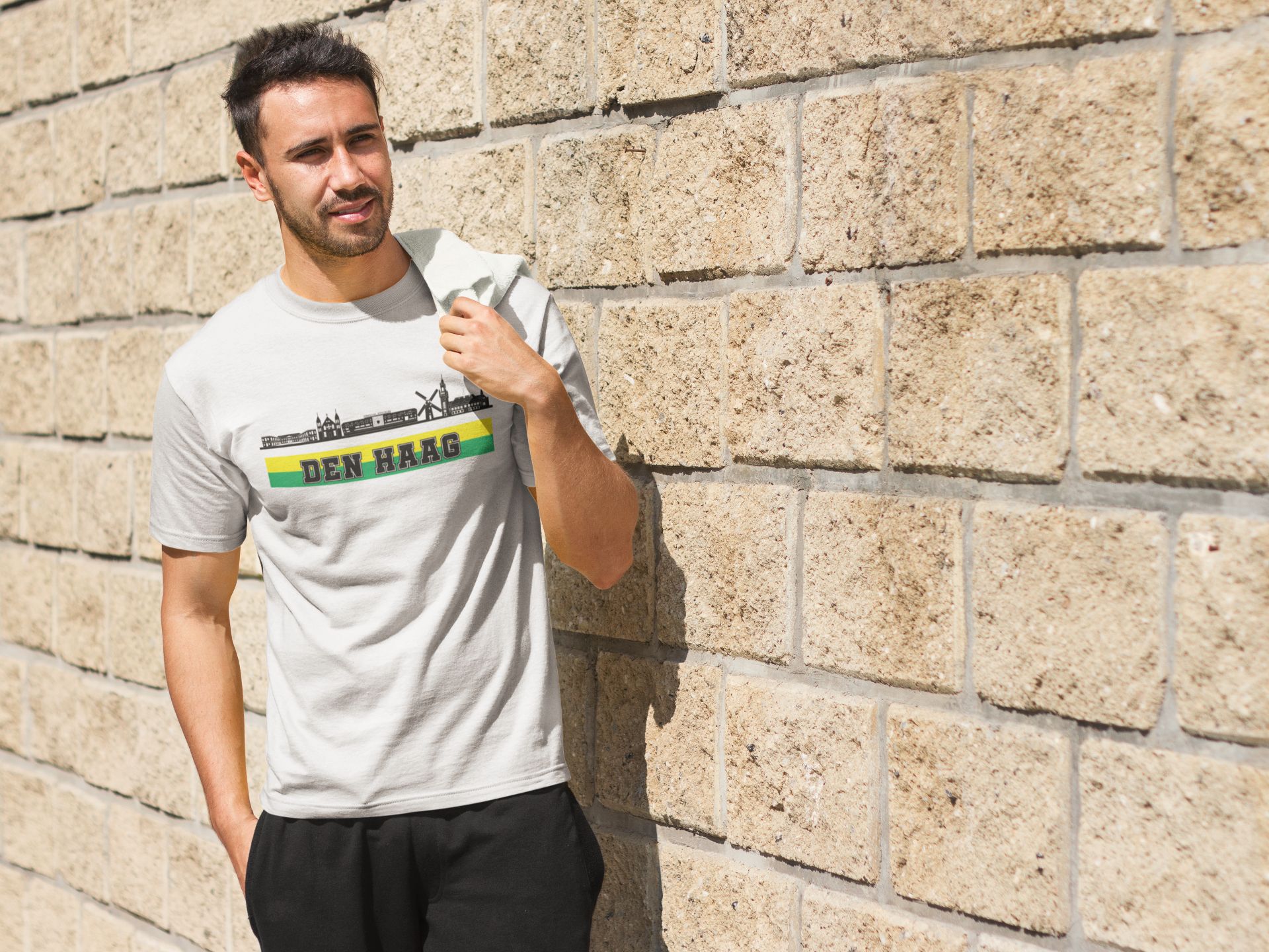 Man draagt een wit T-shirt met de skyline van Den Haag en de tekst “Den Haag” in groen-gele kleuren – ideaal voor trotse Hagenaren en supporters.
