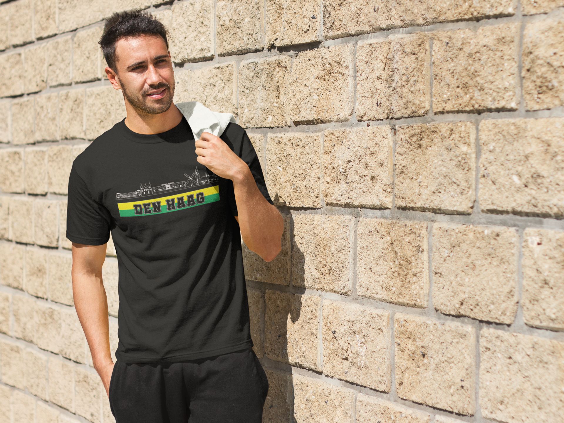 Man draagt een wit T-shirt met de skyline van Den Haag en de tekst “Den Haag” in groen-gele kleuren – ideaal voor trotse Hagenaren en supporters.