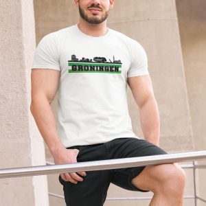 Man draagt T-shirt met skyline van Groningen en groene tekst "Groningen", verkrijgbaar in meerdere kleuren.